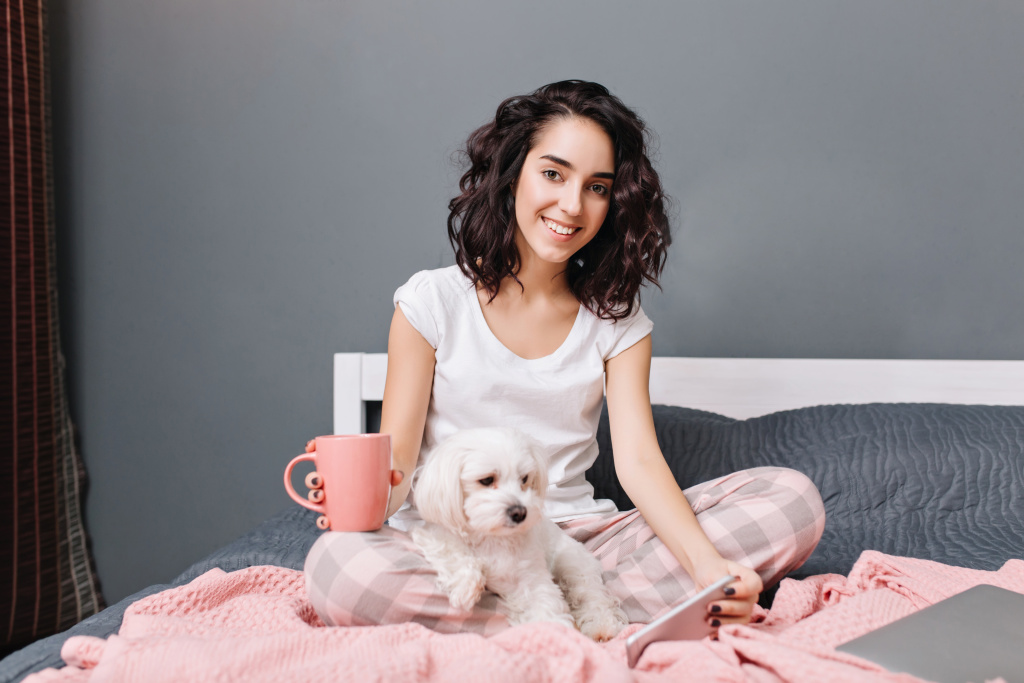 joyful-young-woman-with-curly-brunette-hair-in-pajamas-chilling-on-bed-with-little-dog-in-modern-appartment-pretty-model-relaxing-at-home-with-cup-of-coffee-chatting-on-phone-smiling.jpg
